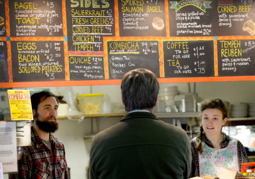 A man and woman stand at a café counter, discussing the menu, which features various food and drink options.
