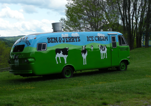 A colorful Ben & Jerry's ice cream truck parked on grass, featuring cow graphics and a blue sky design.