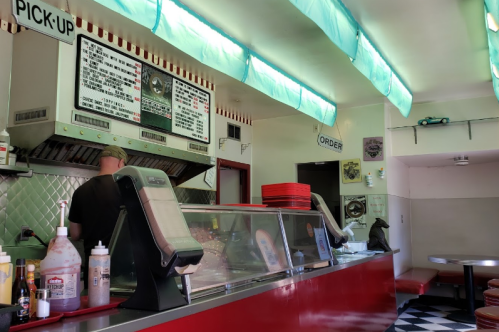 A retro diner interior with a red counter, menu board, and a cook preparing food in a bright, colorful setting.