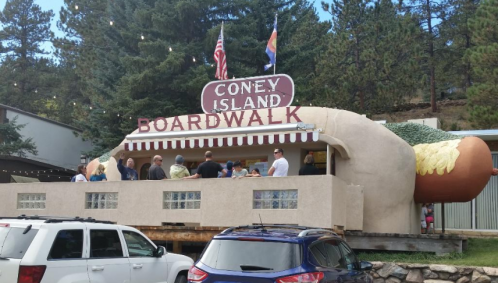 A whimsical building shaped like a hot dog, labeled "Coney Island Boardwalk," with people lined up outside.