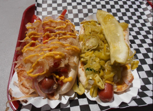 Two hot dogs on a tray, one topped with onions, mustard, and relish, the other with pickles and jalapeños.