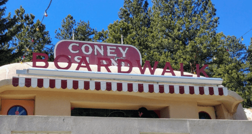 Sign for "Coney Boardwalk" on a building, surrounded by trees and a blue sky.