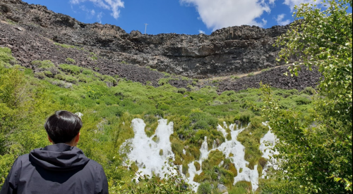 A person stands in front of a lush green landscape with a waterfall, framed by rocky cliffs and a blue sky.