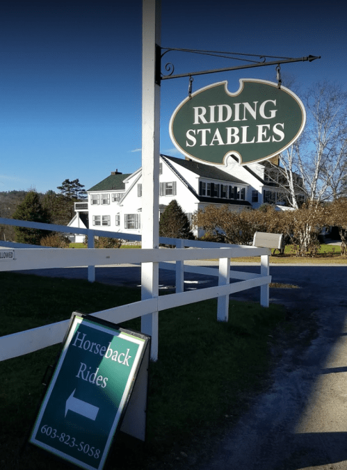 Sign for "Riding Stables" with a direction for "Horseback Rides" and contact number, set in a rural landscape.