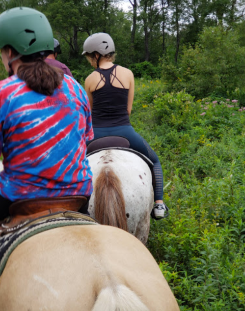 Two people riding horses through a lush, green trail, wearing helmets and casual clothing.