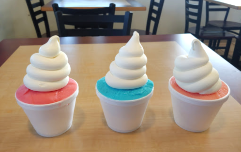Three cups of soft serve ice cream: one pink, one blue, and one red, each topped with a swirl of white ice cream.