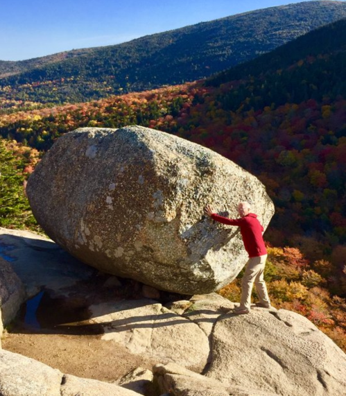 A person in a red sweater leans against a large boulder on a rocky ledge, surrounded by colorful autumn foliage.