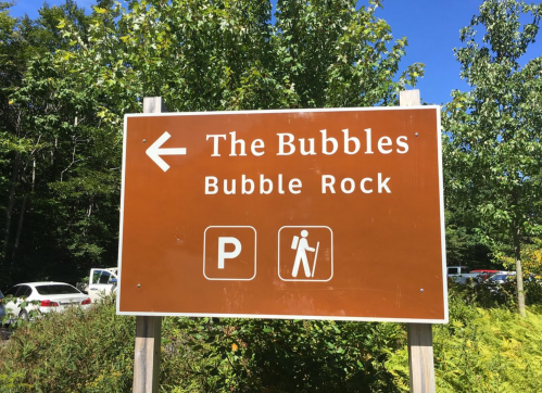 Sign for "The Bubbles" and "Bubble Rock" with directions for parking and hiking, surrounded by trees and blue sky.