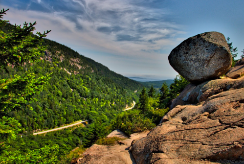 A large boulder sits on a rocky outcrop overlooking a lush green valley and winding road under a blue sky.