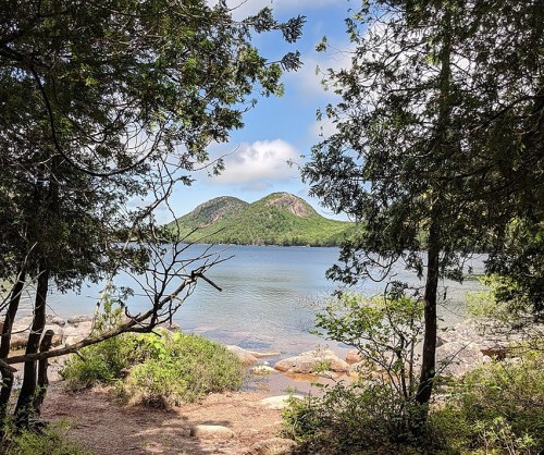 A serene view of a lake framed by trees, with two hills rising in the background under a partly cloudy sky.