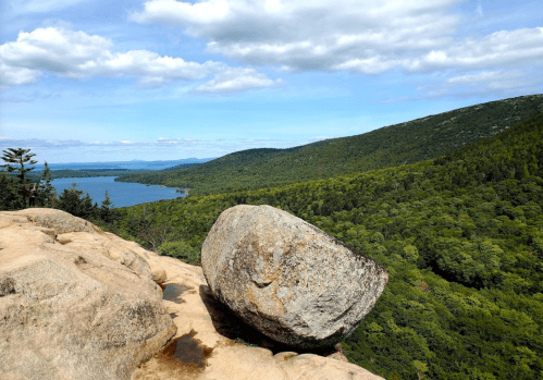 A large boulder sits on a rocky outcrop overlooking a lush green valley and a serene lake under a blue sky.