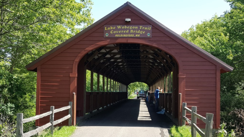 A red covered bridge on the Lake Wobegon Trail, surrounded by greenery and a gravel path.