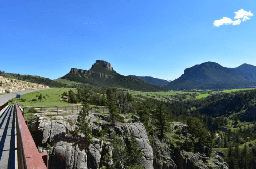 A scenic view of mountains and lush green valleys under a clear blue sky, with a rocky ledge in the foreground.