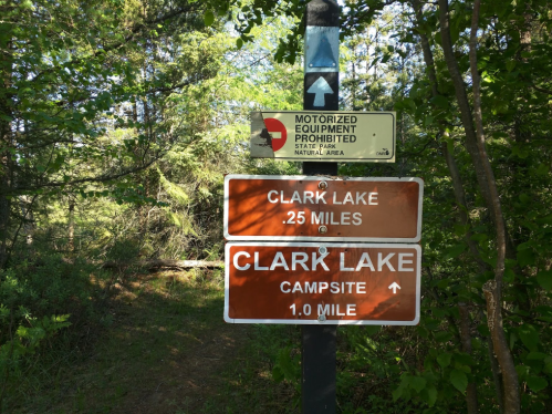 Trail signs indicating directions to Clark Lake, with distances of 25 miles and 1 mile to the campsite.