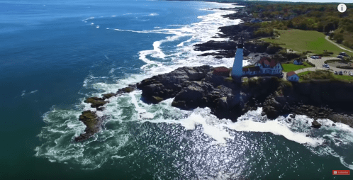 Aerial view of a lighthouse on rocky coast, surrounded by blue ocean waves and a scenic shoreline.
