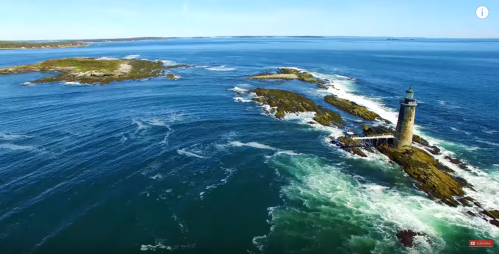 Aerial view of a lighthouse on rocky shores, surrounded by blue ocean and waves under a clear sky.