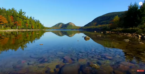 A serene lake reflects mountains and colorful trees under a clear blue sky. Rocks are visible beneath the calm water.