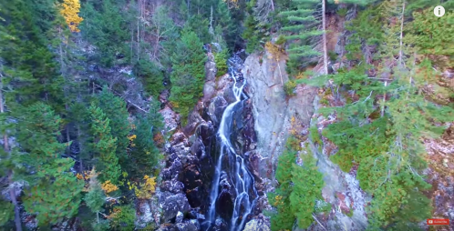 Aerial view of a cascading waterfall surrounded by lush green trees and rocky cliffs in a serene forest setting.