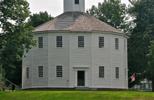 A round white building with a golden cupola, surrounded by trees and grass, featuring large windows and a front door.