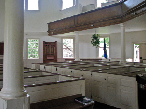 Interior of a historic church featuring wooden pews, tall windows, and a balcony with a hanging plant.