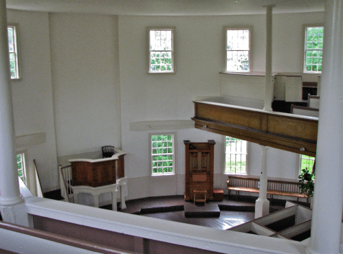Interior view of a simple, bright church with wooden benches, a pulpit, and large windows letting in natural light.