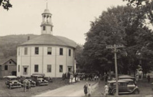 Historic round building with a cupola, surrounded by trees and vintage cars, with people gathered outside.