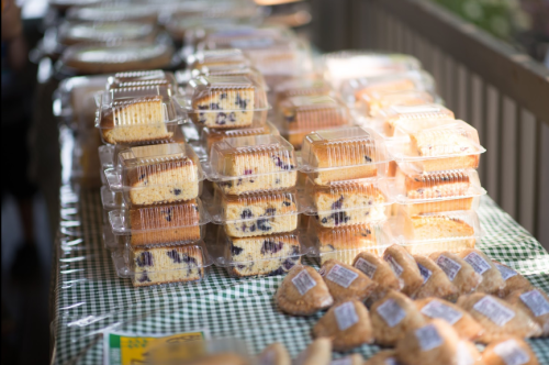 Stacks of packaged baked goods on a checkered tablecloth at a market, including cakes and cookies.