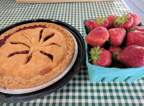 A freshly baked strawberry pie next to a basket of ripe strawberries on a checkered tablecloth.