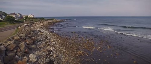 A rocky shoreline meets calm ocean waves under a clear sky, with houses visible along the coast.