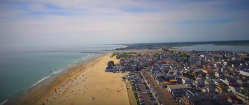 Aerial view of a coastal town with a sandy beach, ocean waves, and a line of houses along the shore.