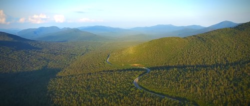 Aerial view of a winding road through lush green mountains and forests under a clear blue sky.