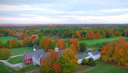 Aerial view of a farm surrounded by vibrant autumn foliage and rolling hills under a cloudy sky.
