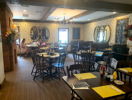 Cozy restaurant interior with wooden tables, yellow placemats, floral wallpaper, and patrons dining in booths.