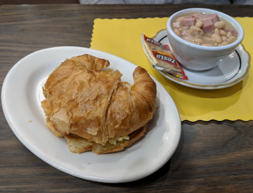 A flaky croissant sandwich on a plate next to a bowl of bean soup with ham, accompanied by a condiment packet.
