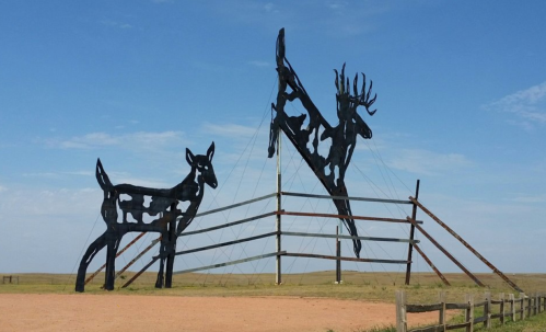Large black silhouettes of a deer and an antelope against a blue sky, set near a wooden fence on a grassy landscape.