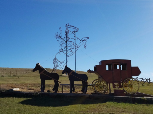 A metal sculpture of a cowboy on horseback next to a stagecoach and two horses against a clear blue sky.