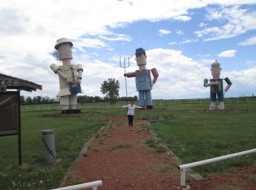 A person stands on a path surrounded by three large, colorful farmer statues in a grassy field under a cloudy sky.