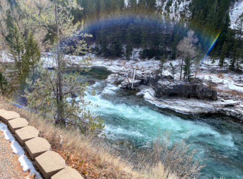 A river flows through a rocky landscape, with a rainbow arching over the water and trees lining the banks.