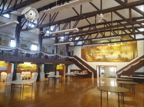 A spacious hall with wooden beams, tables, and murals on the walls, featuring a staircase and chairs arranged for an event.