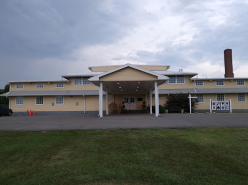 A large, two-story yellow building with a covered entrance and a grassy area in front, under a cloudy sky.