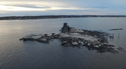 Aerial view of a rocky shoreline with a small, weathered structure on an island, surrounded by calm water and a cloudy sky.