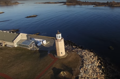 Aerial view of a lighthouse by the water, with rocky shoreline and a building nearby, under a clear sky.
