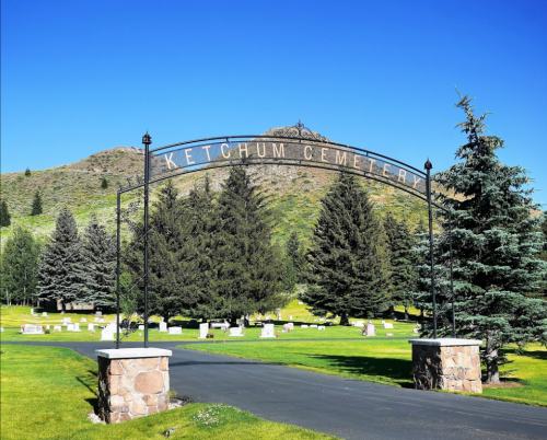 Entrance to Ketchum Cemetery, featuring a metal archway and surrounded by trees and gravestones under a clear blue sky.