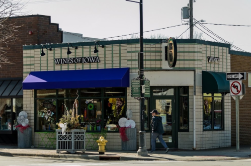 A corner store labeled "Wines of Iowa" with a blue awning, featuring decorative elements and a pedestrian walking by.