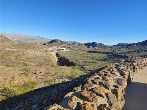 A rocky landscape with mountains in the background under a clear blue sky, featuring a stone wall in the foreground.