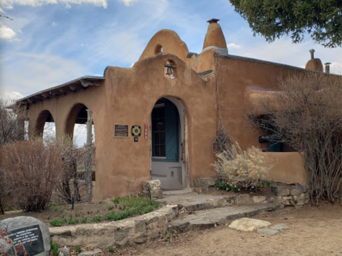 Adobe-style building with arched entrance, surrounded by sparse vegetation and stone pathway under a blue sky.