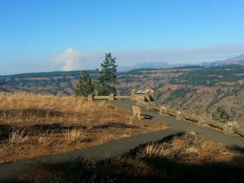 A scenic overlook with a path, benches, and distant mountains under a clear blue sky, with smoke rising in the background.