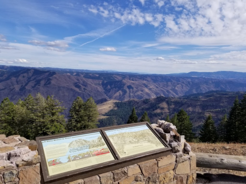 Scenic mountain view with informational signs in the foreground, surrounded by trees and a blue sky with clouds.