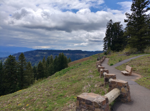 A scenic view of a mountain path lined with stone benches, surrounded by trees and under a cloudy sky.