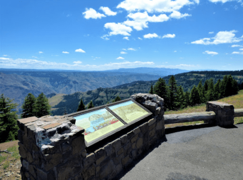 Scenic overlook with informational signs, surrounded by lush trees and mountains under a clear blue sky.